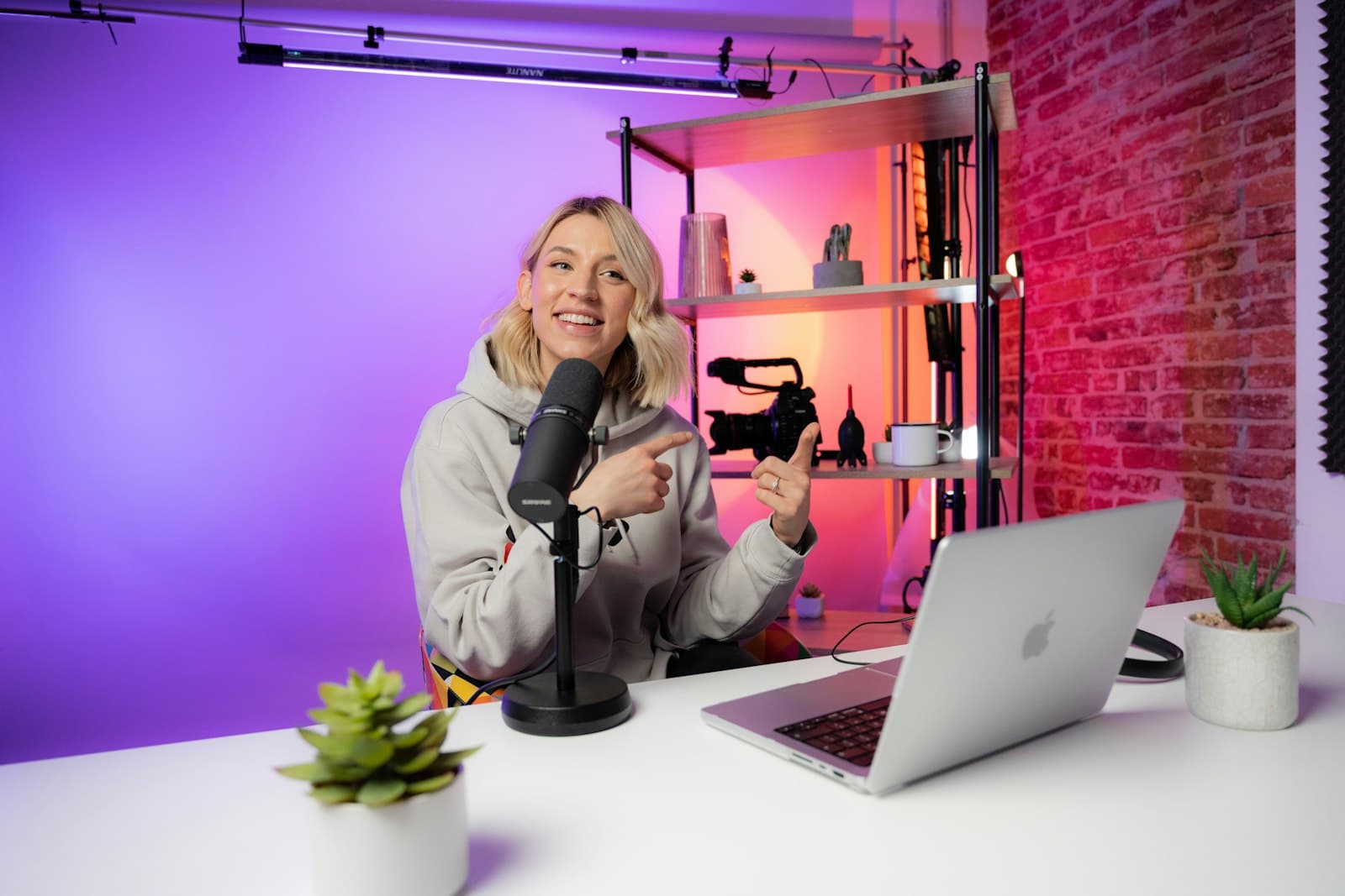 Smiling woman creator at a desk with a microphone and laptop in a studio lit purple and pink